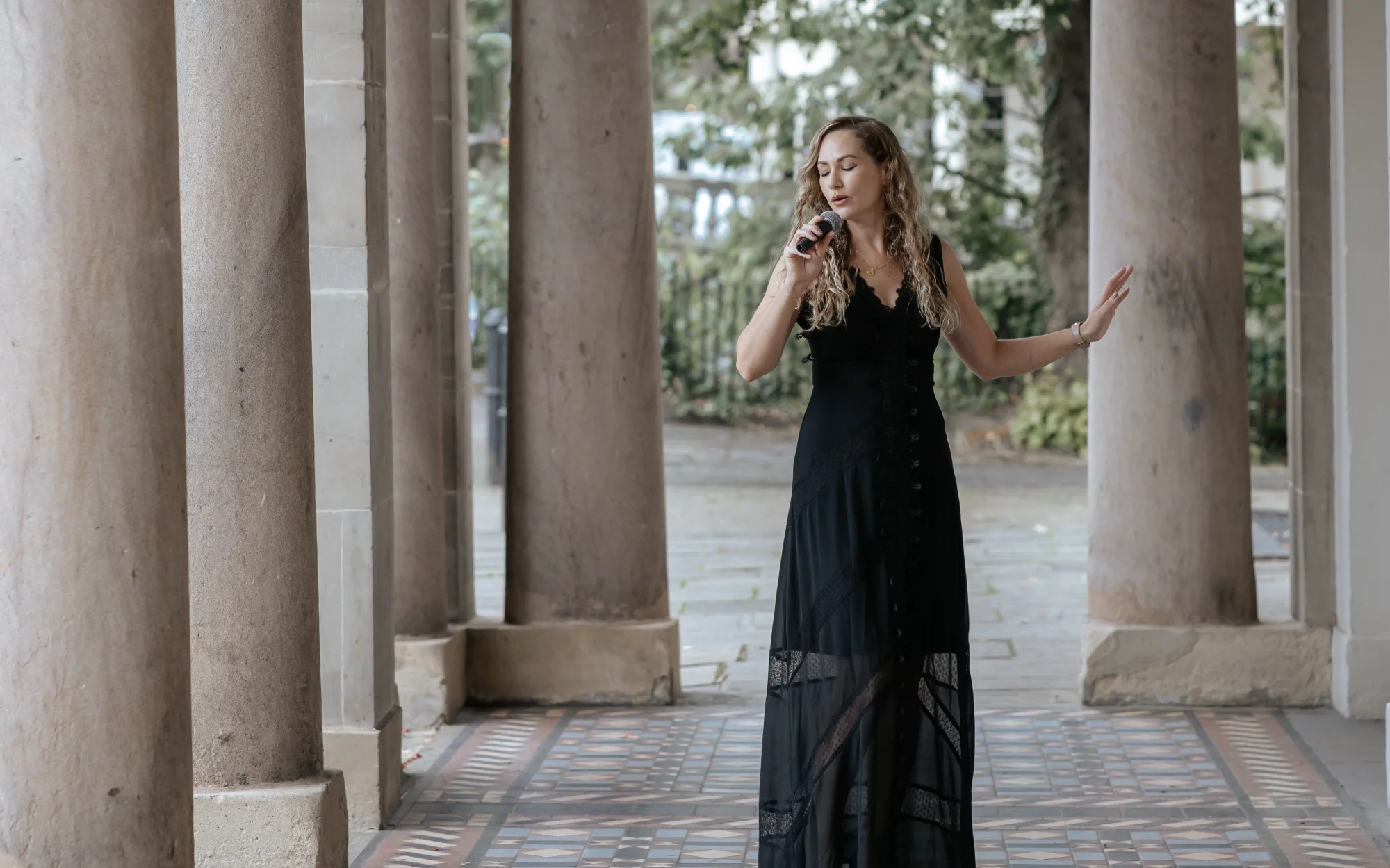 Wedding & event singer Songbird Gebby in dark dress singing into microphone, surounded by Doric columns.